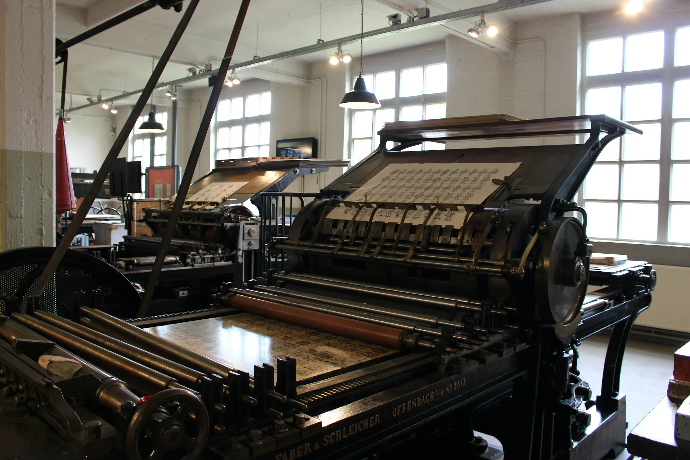 A Faber & Schleicher flatbed cylinder press in a bright, white-walled print workshop, a sheet of patterned proofs resting on its feed board